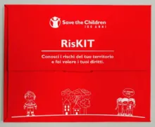 Red box with the Save the Children logo and the title “RisKIT.” The subtitle reads: “Know the risks in your area and claim your rights.” The design includes simple white line drawings of a child, houses, trees, and a group of people, with one speech bubble saying “I think so!”